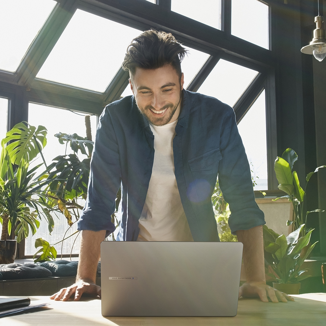 一名男性员工正微笑看着 ASUS ExpertBook 笔记本电脑。他站在桌前，他身后有一些绿植